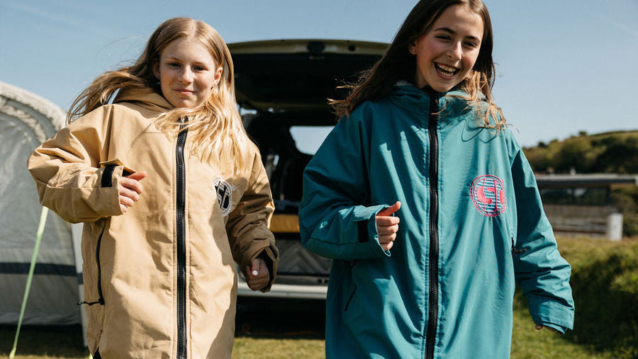 Two people wearing long raincoats walking outdoors with a tent and vehicle in the background.