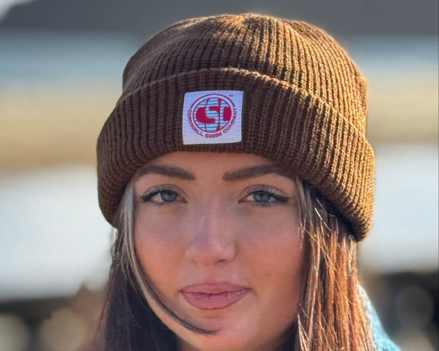 Woman wearing a brown beanie with a logo, standing outdoors.